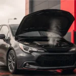 Dark grey car with its bonnet open and light steam rising from the engine bay, indicating a possible burning smell or overheating issue.